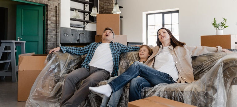 A couple relaxes on a plastic-covered couch amidst moving boxes in a modern, spacious living room.