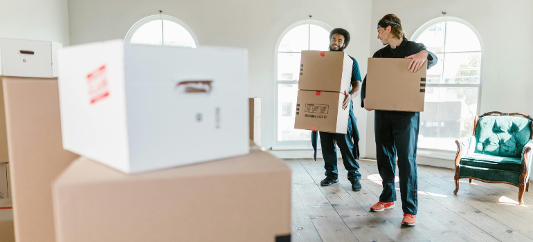 Two movers carry stacked boxes in a spacious, bright room with large windows and a vintage green armchair in the background.