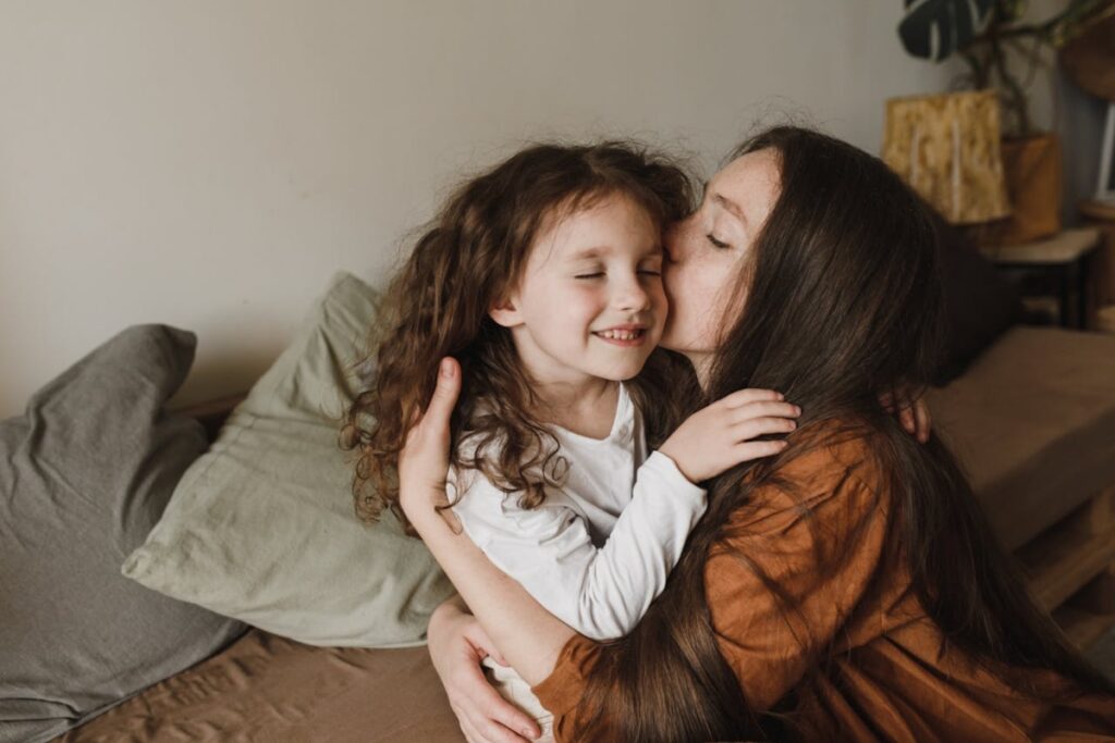 A woman with long brown hair embraces a child with curly hair, sitting on a couch surrounded by soft pillows in a cozy setting. Children need support before a move across the country