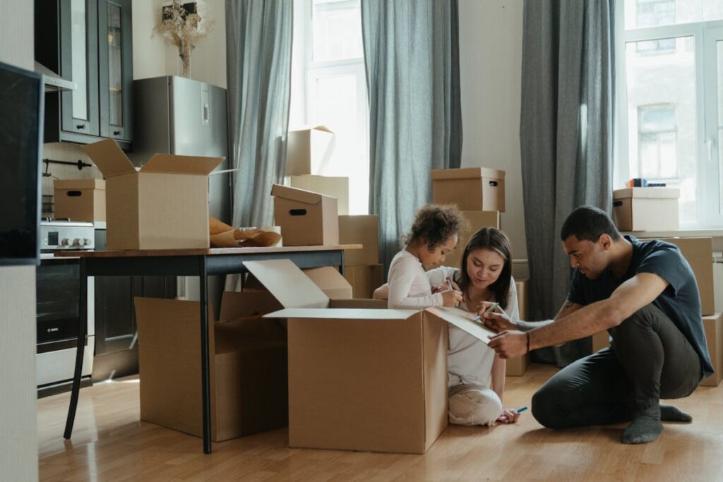 A family is unpacking boxes in a cozy kitchen, with children drawing on cardboard with their parent nearby.