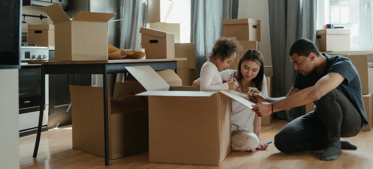 A family is unpacking cardboard boxes in a cozy living room, surrounded by more boxes, with one adult assisting a child.