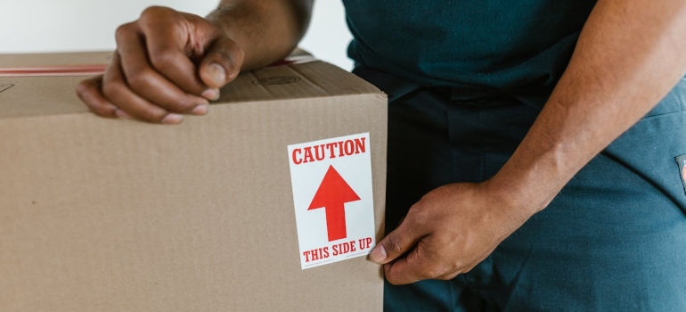 A person placing caution label on a cardboard box, indicating the correct orientation with an arrow pointing up.
