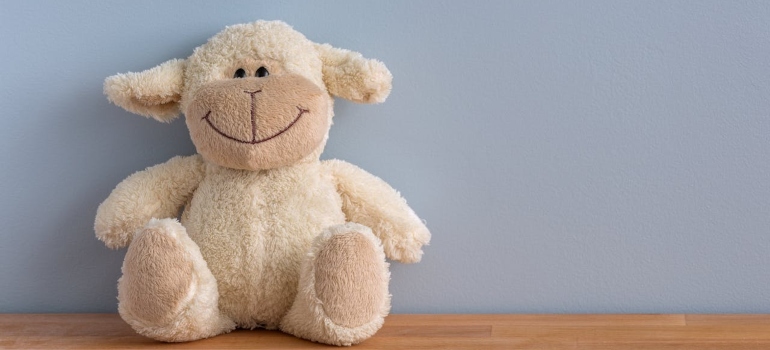 A plush, smiling sheep toy sits on a wooden surface against a light blue wall, exuding warmth and friendliness. Toys bring support for children moving across the country.